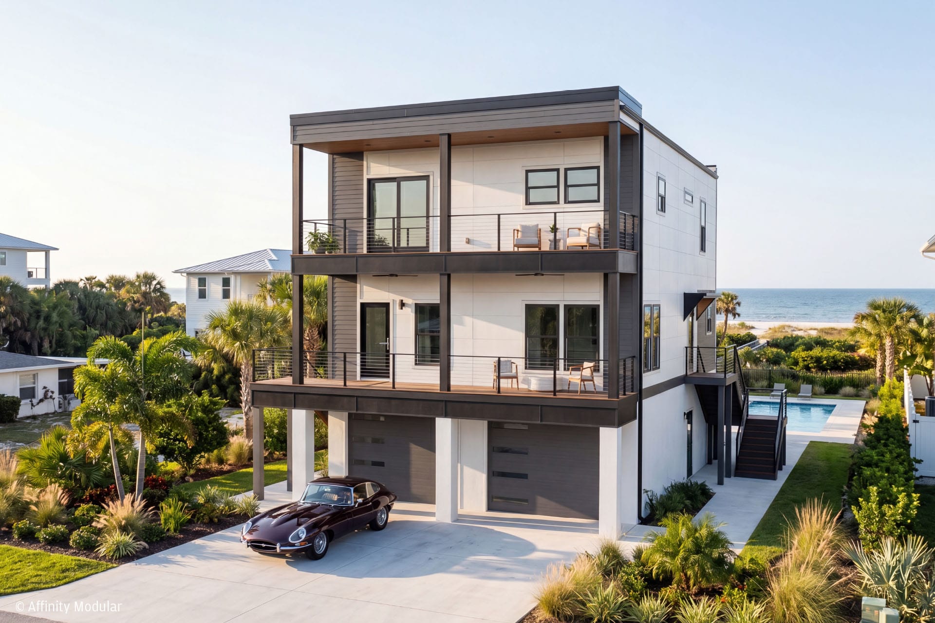 Modern three-story modular home with balconies, a pool, and a classic car parked in the driveway by the ocean.