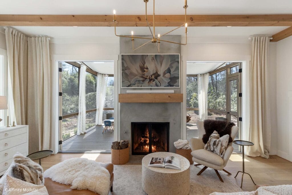 Modern modular home living room with a fireplace, cozy seating, and large doors opening to a screened porch with curtains.