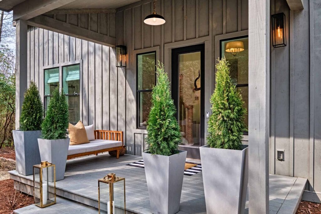 Modern front porch with gray siding, potted evergreens, a wooden bench, and lanterns on tiled floor.