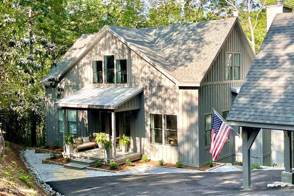 Gray modern farmhouse modular home with front porch, American flag, greenery, and sloped driveway surrounded by trees.