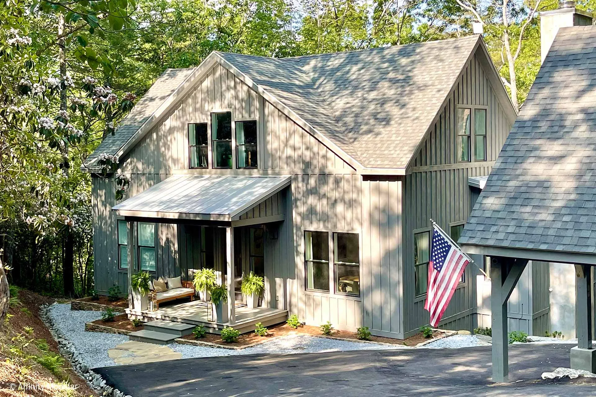 Gray modern farmhouse modular home with front porch, American flag, greenery, and sloped driveway surrounded by trees.