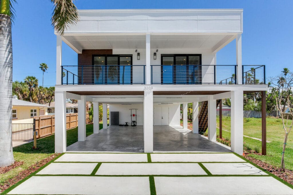 Modern elevated white modular home with open parking area beneath and balcony, surrounded by greenery and palm trees.