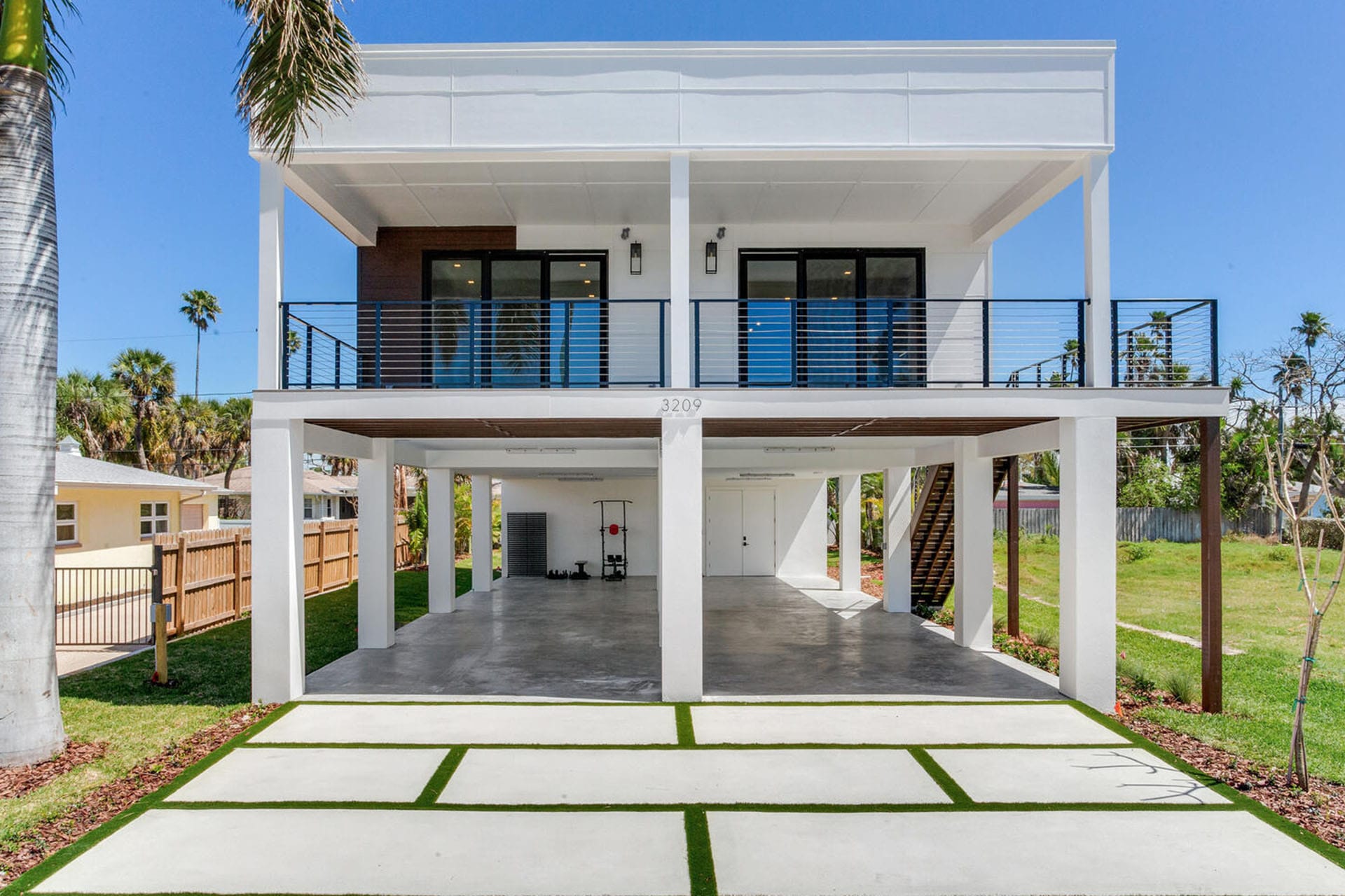 Modern elevated white modular home with open parking area beneath and balcony, surrounded by greenery and palm trees.