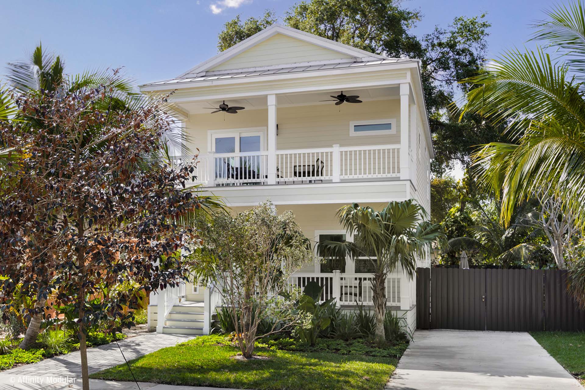 Two-story white modular home with front porches, surrounded by palm trees and lush greenery on a sunny day.