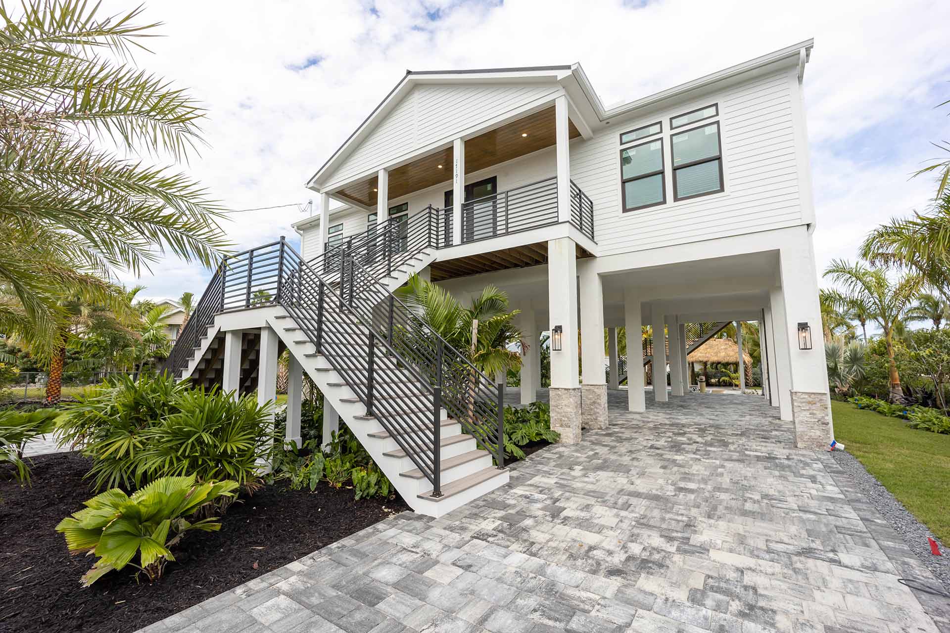 Elevated modular home with large front stairs, palm trees, and a paved driveway under a partly cloudy sky.