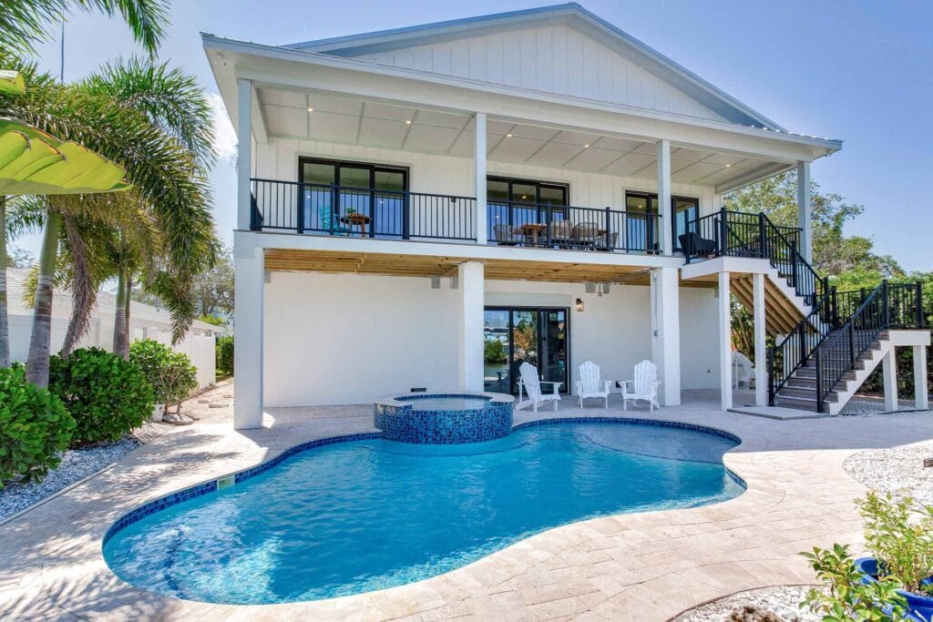 Two-story modular home with balcony, pool, hot tub, palm trees, and patio chairs in a sunny backyard setting.