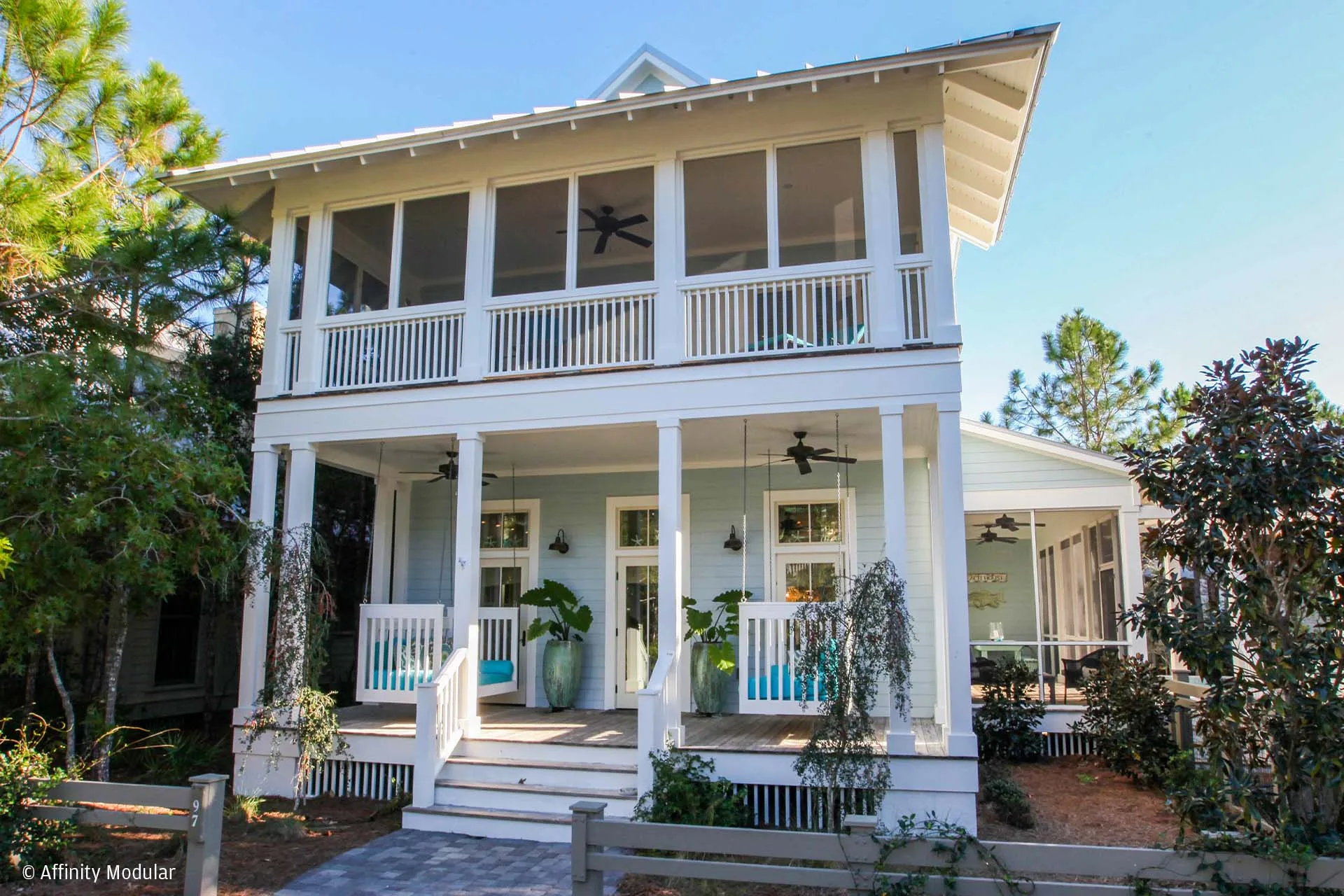 Two-story white modular home with a large front porch, balcony, columns, and surrounding greenery on a sunny day.