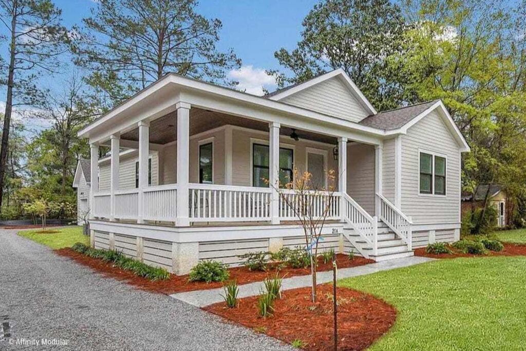 A charming single-story modular home with a wide front porch, white railings, and a neatly landscaped yard.
