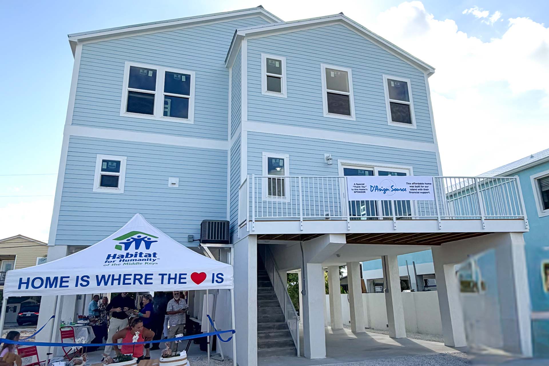 Two-story blue modular home with people gathered under a Habitat for Humanity tent.
