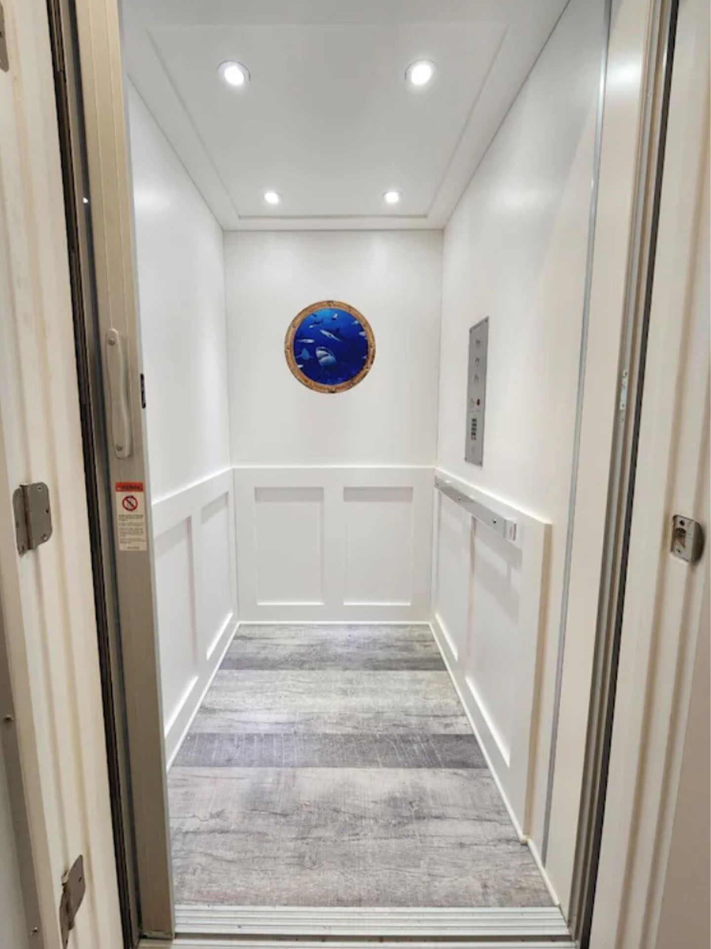 A small, modern white elevator with wood-patterned flooring and a round blue wall art at the back, perfect for any modular home.